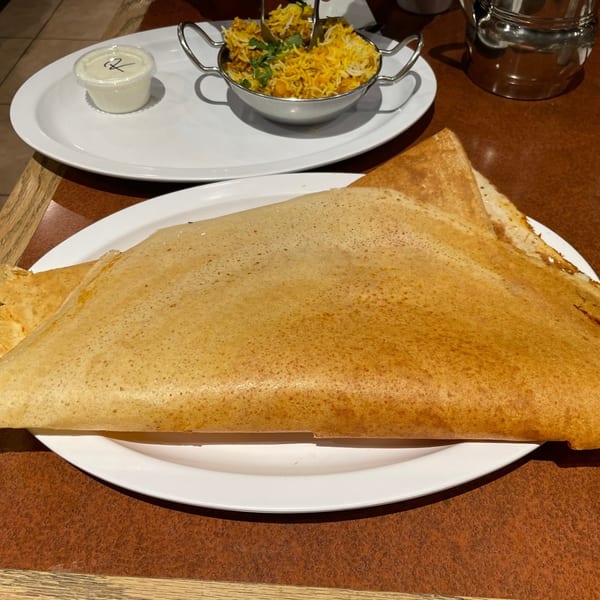 A large golden-brown dosa on a white plate, with a side of biryani and sauce.